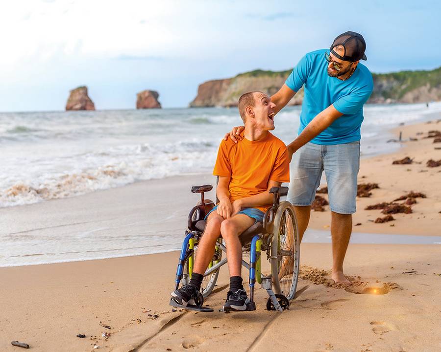 Junger Mann mit Behinderung im Rollstuhl mit Begleitung am Meer