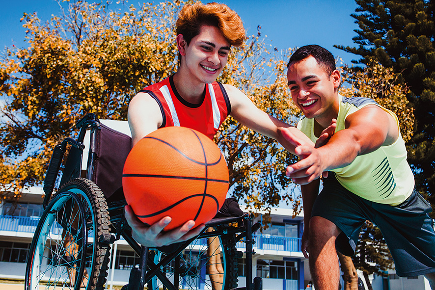 Jugendlicher im Rolllstuhl spielt mit einem Freund Basketball im Freien