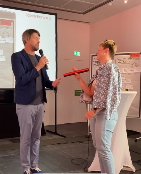 Das Foto zeigt die Staffelstabübergabe des Moderators an eine Teilnehmerin aus Forum 2, die Ideen im Plenum präsentiert hatte.